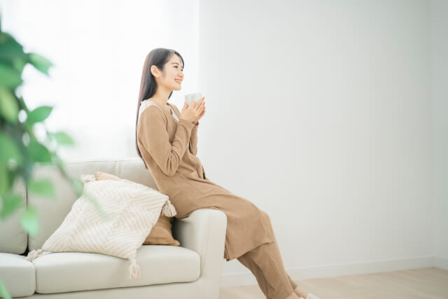 白いソファに腰掛けて温かい飲み物を飲んでいる女性の画像